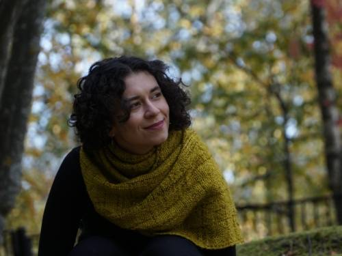 Eine Frau mit einem gelben Schal sitzt lächelnd vor einem Zaun.