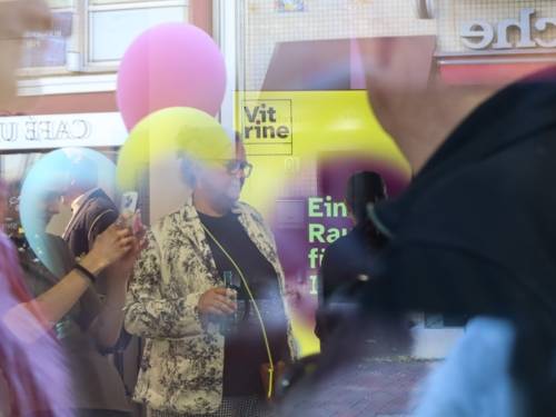 Mehrere Menschen, einige Luftballons und ein Aufsteller. Fotografiert durch eine Glasscheibe. Formen und Farben verschwimmen leicht und ergeben eine Art Collage. Die Farben gelb und lila dominieren das Bild.