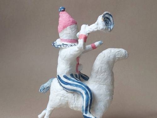 Zu sehen ist eine kleine Figur gefertigt von Kira Kotliar. Auf einem weißen vierbeinigem Tier sitzt ein kleines Männchen und bläst in eine Tröte.