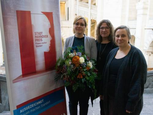 Drei Frauen stehen neben einem Banner mit der Aufschrift "Stadt-Hannover-Preis, Frauen machen Standort", eine der Frauen hält einen großen Blumenstrauß.