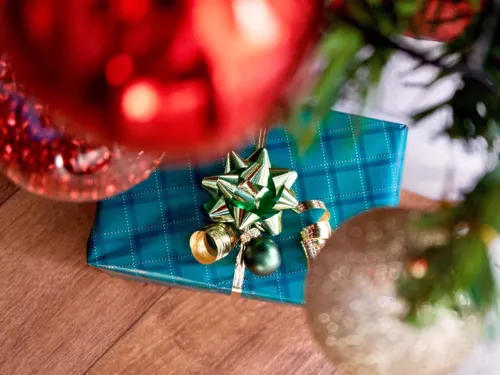 Ein Geschenk liegt unter dem Weihnachtsbaum, im Vordergrund hängen Christbaumkugeln