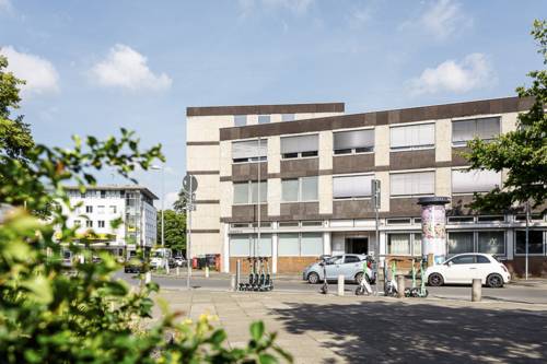 Ein Bild des Gebäudes in der Leinstraße 14. Im Vordergrund links ein wenig Pflanzengrün, in der Bildmitte das Gebäude mit dem Haupteingang, davor parkende Autos und E-Roller.