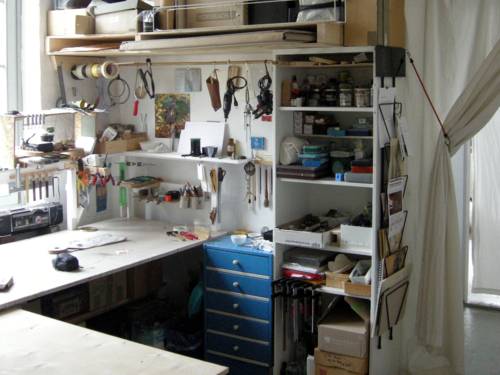 Blick in das Atelier eines Künstlers: Eine Arbeitsfläche in L-Form ist umgeben von allerhand künstlerischem Material und Werkzeug.