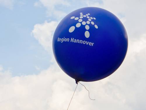 Ein blauer Luftballon schwebt vor einem bewölkten Himmel in der Luft, auf dem Ballon ist in weißer Farbe das Logo der Region Hannover.