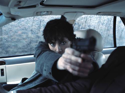 Eine Person sitzt auf dem Beifahrersitz eines Autos und zielt mit einer Waffe auf jemanden, der nicht im Bild zu sehen ist.