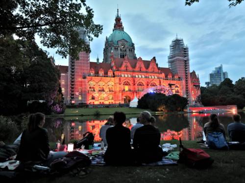 Menschen, die abends in einem Park sitzen. Sie blicken auf eine beleuchtete Bühne vor dem Neuen Rathaus.