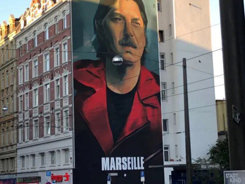 Sichtbar ist ein H&auml;userblock dessen Seite mit einem Portraitbild eines Mannes bemalt ist. Das Portrait stellt den Charakter "Marseille" aus der Serie Haus des Geldes dar.
