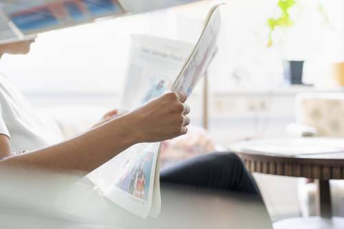 Eine Person in weißem Shirt ließt eine aufgeschlagene Zeitung. Von der Person erkennt man nur die Unterarme und das Kinn.
