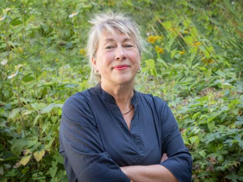 Eine Frau mit blonden Haaren steht vor grünen Pflanzen. Sie hat verschränkte Arme, der Gesichtsausdruck ist freundlich aber bestimmt.