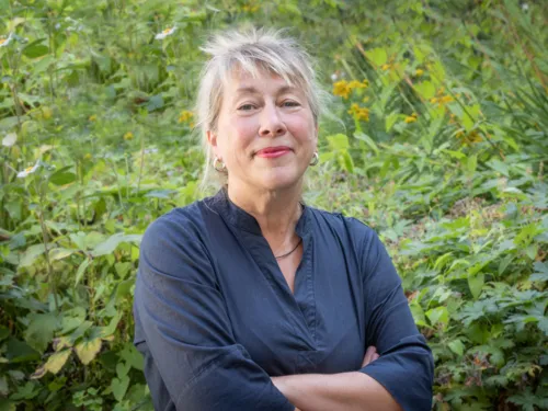 Eine Frau mit blonden Haaren steht vor grünen Pflanzen. Sie hat verschränkte Arme, der Gesichtsausdruck ist freundlich aber bestimmt.