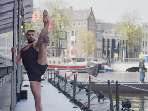 Das Bild zeigt eine Person in schwarzer Tanzkleidung, die auf einer Bühne am Wasser eine anspruchsvolle Dehn- und Ballettpose ausführt. Im Hintergrund sind ein Kanal, Boote und historische Gebäude einer Stadt zu sehen. Das weiche Tageslicht und die urbane Kulisse bilden einen spannenden Kontrast zur eleganten Bewegung und Körperbeherrschung der dargestellten Person.