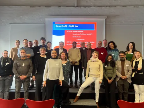 Ein Gruppenbild, auf dem 23 Menschen unterschiedlichen Alters und Geschlechts vor und auf einem kleinen Podest für die Kamera posieren. Im Hintergrund eine Leinwand mit einer Powerpoint-Folie in blau und rot.