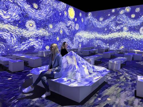 Zu sehen ist ein Raum der immersiven Van Gogh Ausstellung: Das Gemälde „Die Sternennacht“ bespielt Projektionen den hellen und mit Sitzmöbeln ausgestatteten Raum.