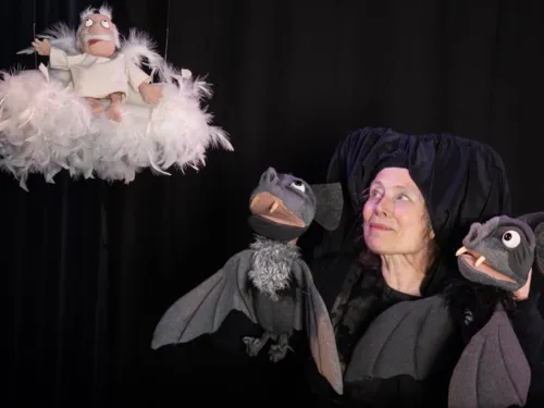 Dunkel gekleidete Puppenspielerin mit zwei Fledermaus-Handpuppen und einer Puppe, die eine älteren Mann als Engel auf einer Wolke darstellt.