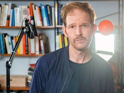 Mann mit kurzem Haar und Bart, im Hintergrund eine Schreibtischlampe und ein Bücherregal