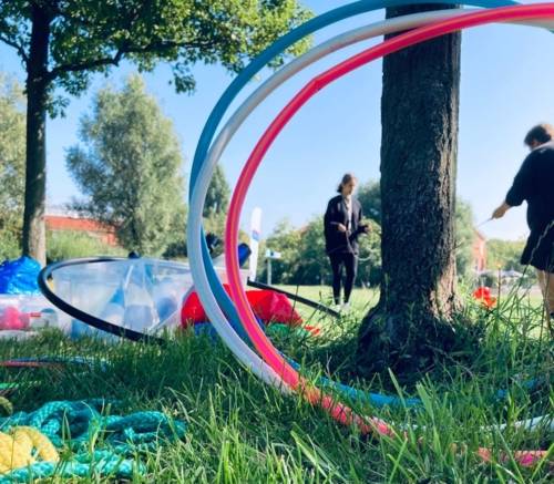 Hula Hoop reifen stehen an einen Baum gelehnt, daneben liegen Keulen und andere Zirkusutensilien. Im Hintergrund spielen Menschen mit einem Diabolo.