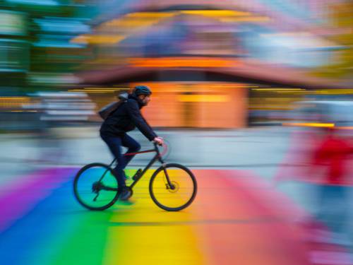 Zu sehen ist eine Fotografie von Amlinger Full: Ein Fahrradfahrer im Profil vor einem verschwommenen, bunten Stadtbild.