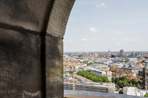 Ein Blick über die Dächer von Hannover. Sonniger Tag. Im linken Drittel des Bildes ein Ausschnitt eines dunklen Sandsteinbogens.