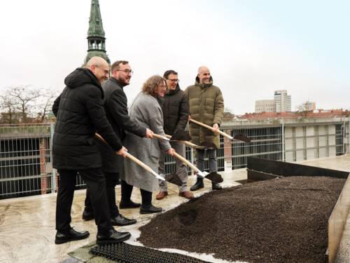 Fünf Personen stehen auf einem Dach vor einem Beet und halten Spaten mit Erde vor sich, hinter ihnen sind Dächer und ein Kirchturm zu sehen.