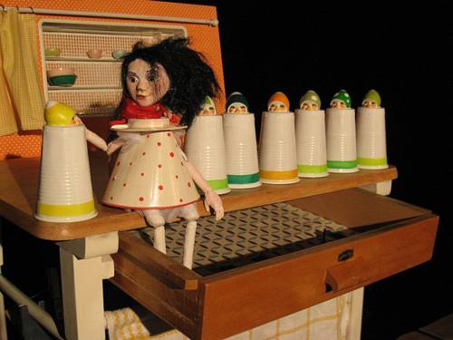 Zu sehen ist eine Puppe, die auf einem Schrank sitzt. Die Schublade unter ihr ist geöffnet.