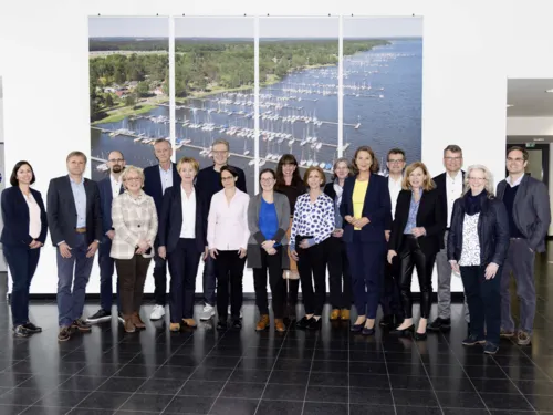 Gruppe von Personen vor einem Bild auf dem ein Gew&auml;sser, Bootsanleger und Segelboote zu sehen sind.
