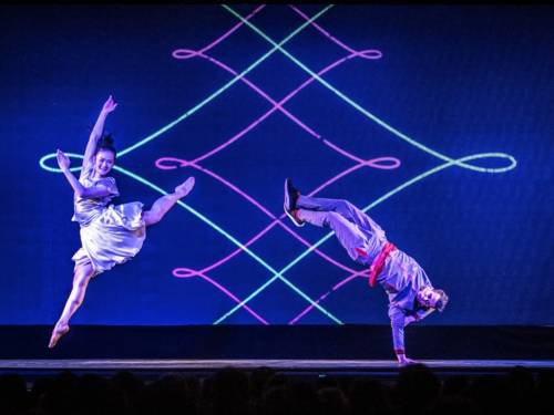 Zu sehen sind eine Akrobatin und ein Akrobat, die vor einem Neonmuster auf Leinwand eine Tanzperformance vorführen.