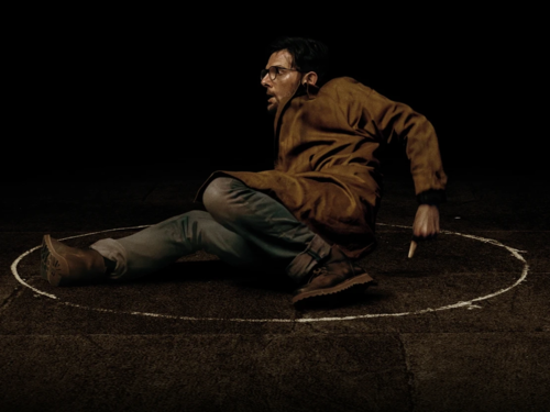 Das Bild zeigt eine einzelne Person auf einer dunklen Bühne oder in einem schwach beleuchteten Raum. Die Person sitzt auf dem Boden innerhalb eines mit Kreide gezeichneten Kreises und hält ein Kreidestück in der Hand. Die Kleidung besteht aus einem braunen Mantel, heller Hose und dunklen Schuhen. Der schwarze Hintergrund und die gezielte Beleuchtung lenken den Blick auf die Person und den markierten Kreis, wodurch eine reduzierte und konzentrierte Bildwirkung entsteht.
