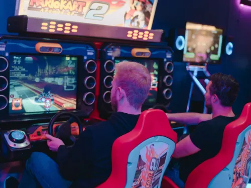 Zwei Männer sitzen in roten Arcade-Rennsitzen und spielen „Mario Kart Arcade GP 2“ in einer Spielhalle.