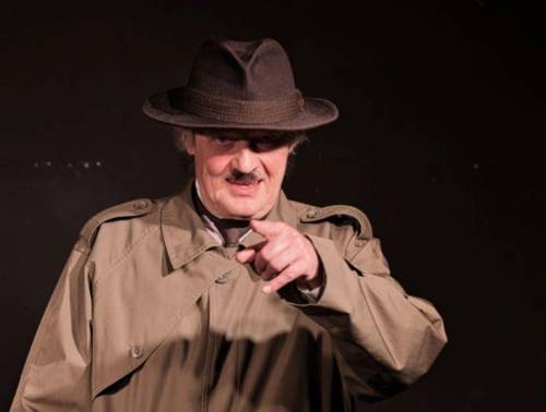 Mann mit Trenchcoat und Hut deutet mit dem linken Zeigefinger in die Kamera.