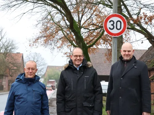 Drei Männer stehen auf dem Bürgersteig neben einer Straße, direkt hinter ihnen ist ein Schild "Tempo 30".