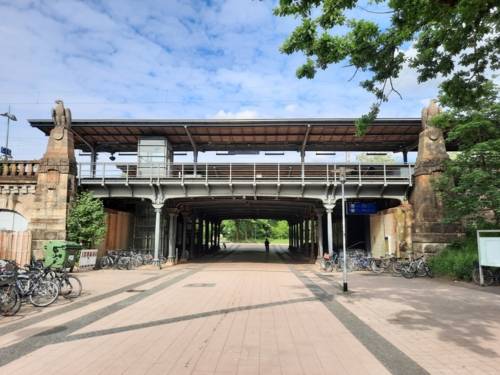 Ein Weg führt unter einer Brücke mit Stahlträgern und seitlichen Steinsäulen hindurch, darüber ein Bahnsteig mit Dach, der durch ein transparentes Geländer zu sehen ist.