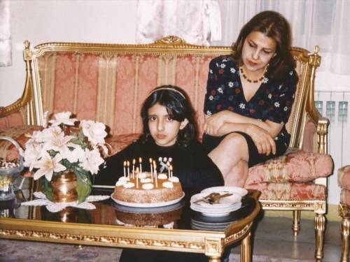 Zu sehen ist ein altes Geburtstagsfoto: Das Geburtsratskind sitzt auf dem Boden, vor ihm steht eine Torte auf einem Couchtisch. Hinter ihr sitzt eine Frau auf einer Couch und schaut schräg von oben auf das Kind.