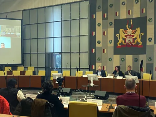 Bild aus dem Ratssaal der LHH. Im Vordergrund drei Menschen, die mit dem Rücken zur Kamera an Plenartischen sitzen. Bildmitte rechts drei Personen, die der ersten Gruppe gegenübersitzen. Im Hintergrund ein großer Wandteppich mit dem Wappen der Landeshauptstadt Hannover.