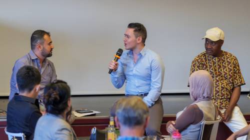 Drei männlich gelesene Personen sitzen auf dem Rand einer Bühne. Die Person in der Mitte spricht in ein Mikrofon und ist der Person links zugewandt. Im Hintergrund eine weiße Leinwand, im Vordergrund sind die Hinterköpfe von mehreren ZuschauerInnen zu sehen.