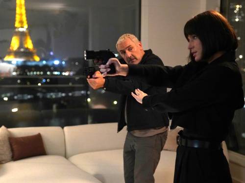 Ein Mann und eine Frau richten ihre Pistolen auf jemanden außerhalb des Bildes. Im Hintergrund der beleuchtete Eiffelturm