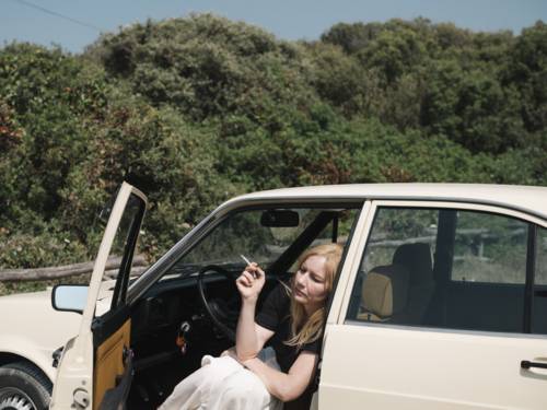Das Bild zeigt eine Person, die in der geöffneten Fahrertür eines hellen Pkw sitzt. Die Person hält eine Zigarette in der Hand und lehnt sich aus dem Fahrzeug heraus. Im Hintergrund erstreckt sich eine dicht bewachsene Landschaft mit Büschen und Bäumen unter klarem Tageslicht. Die natürliche Umgebung, das helle Fahrzeug und die ruhige Komposition prägen die Szene.