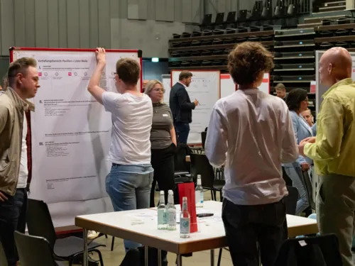 Menschen stehen in einem Raum an Tischen und Stellewänden, ein Mann schreibt etwas auf ein Papier an einer Stellwand.