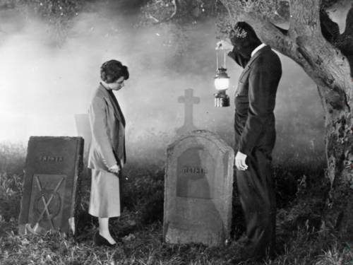 Schwarz‑weiß Szene auf einem nebeligen Friedhof: Zwei Personen stehen sich gegenüber, eine hält eine Laterne. Im Vordergrund sind Grabsteine mit den Aufschriften „FATHER“ und „MOTHER“ sichtbar, im Hintergrund ein Kreuz und ein großer Baum. Die Szene wirkt inszeniert und atmosphärisch dicht.