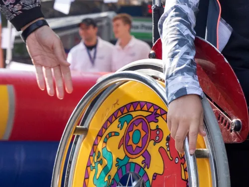 Eine j&uuml;ngere Person f&auml;hrt mit einem Rollstuhl, die Hand einer weiteren Person ist daneben.