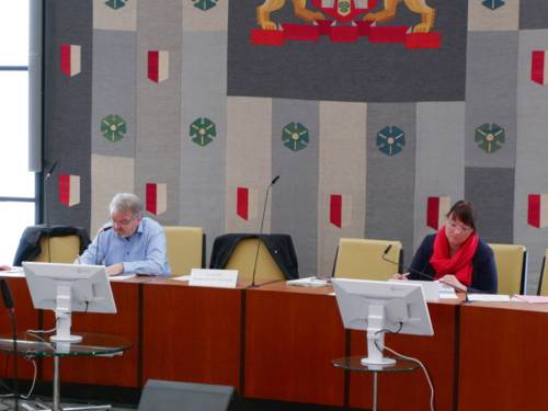 Ein Mann und eine Frau sitzen in einem Planarsaal. Zwischen ihnen sind zwei Stühle frei, vor ihnen steht jeweils ein Monitor. Im Hintergrund hängt ein Wandteppich mit Symbolen und Farben der Landeshauptstadt Hannover.
