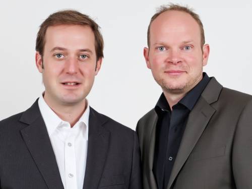 Zwei Männer in Hemd und Sakko, einer mit kurzen Haar, der andere mit Stirnglatze und Fünftagebart.