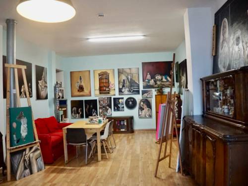 Blick in das Atelier eines Künstlers: Werke hängen an den Wänden, Arbeitsmaterialien wie Pinsel und Staffeleien sind im Raum verteilt und es gibt Möbel wie ein Sofa und einen Schrank mit Malutensilien.