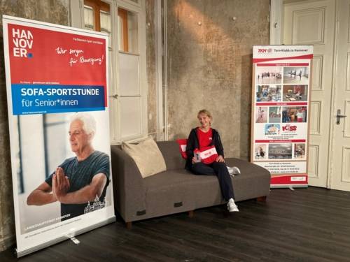 Eine Frau auf einem Sofa. Daneben ein Aufsteller zur Sofa-Sportstunde.