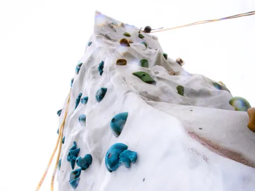 Blick von unten an eine freistehende Outdoor-Kletterwand mit bunten Klettergriffen und Sicherungsseilen.