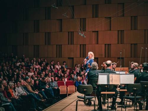 Frau mit Mikrophon neben einem klassischen Orchester vor Publikum.