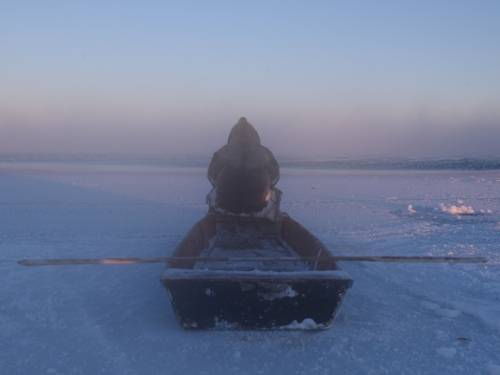 Zu sehen ist eine Fotografie einer Eislandschaft bei Sonnenuntergang. Auf einem Boot sitzt eine Person in dicker Winterkleidung mit dem Rücken zum Betrachter.
