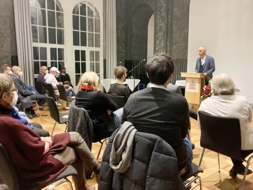 Menschen sitzen in einem Raum, vor ihnen ein Redner am Pult.