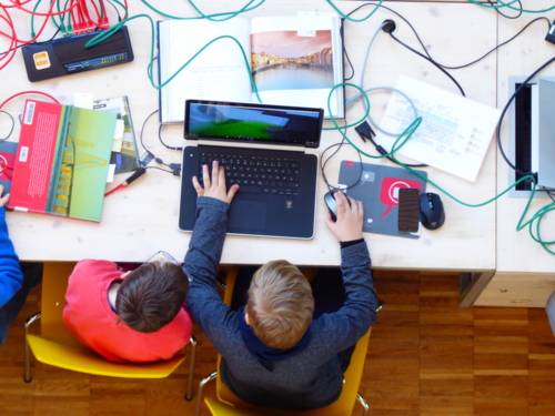 Zwei Kinder sitzen vor einem Laptop, der von Kabeln und Büchern umringt ist.