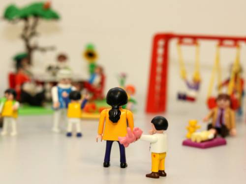 Spielplatzszene mit Spielfiguren: Ein Kind wendet sich an seine Mutter, diese wendet dem Kind den Rücken zu und reagiert nicht. Der Hintergrund ist verschwommen. Dort interagieren andere Eltern mit ihren Kindern.