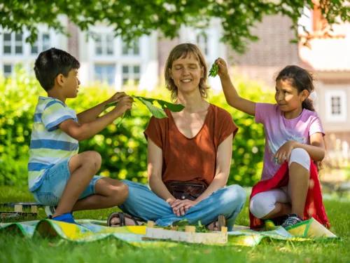 Eine Personen sitzt im Grünen und lauscht den Geräuschen von Blättern, mit denen zwei Kinder ihr zufächern